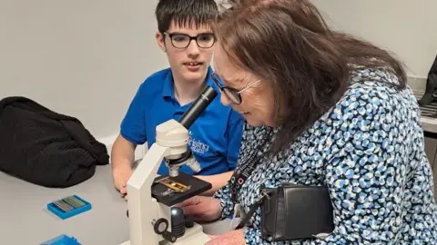 Hull City Council A woman with patterned dress, shoulder length brown hair and glasses looking through a microscope with a boy, who is wearing a blue polo shirt, short brown hair and glasses.
