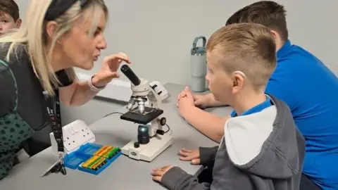 Hull City Council A woman with shoulder-length blonde hair and a headband and black top is using a microscope and looking at a young boy, who has short blonde hair, a grey hoody and dark trousers. He is stood next to a boy with short brown hair, blue top and dark trousers.