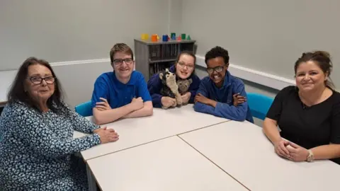 Hull City Council A woman with patterned dress, shoulder length brown hair and glasses sitting at a table with three school pupils wearing blue tops and another woman with brown hair tied back and a black top. They are in a classroom setting.