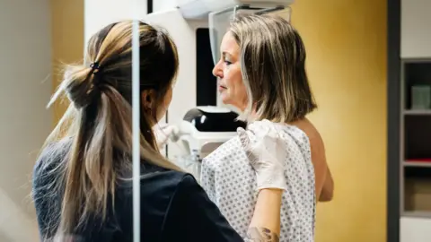 Getty Images A nurse with brown hair half tied back is faced away and providing assistance to a woman with shoulder length hair in a hospital gown who is undergoing a mammogram in a modern medical facility. 