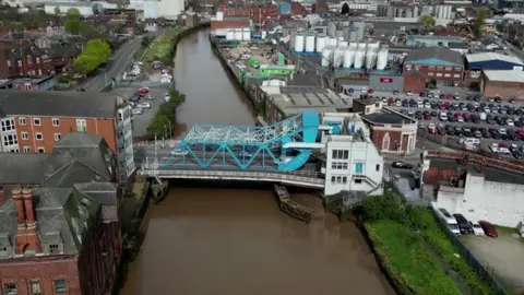 BBC An aerial view of a blue and white bridge spans over a brown river. Industrial looking buildings, tower blocks and car parks are visible on either side of the bank.