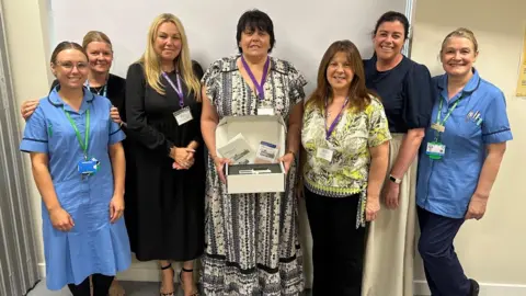 Hull University Teaching Hospitals NHS Trust Hospital staff posing for a picture. Two of the subjects are wearing blue uniforms. A woman in the centre of the image is holding a box containing equipment.