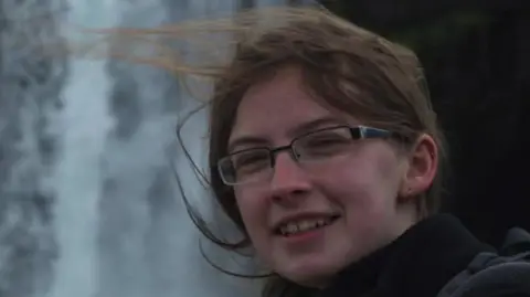 Family Handout Headshot picture of a woman wearing glasses and brown hair who is smiling. Her hair is tied back but is windswept. She is stood outside and is wearing a black coat.