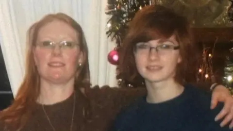 Family handout Woman on the left is wearing glasses and has long ginger hair. She is also wearing a brown jumper with a long gold necklace around her neck. She has her arm around a younger woman who is wearing glasses and short ginger hair in a bob and is wearing a blue jumper.