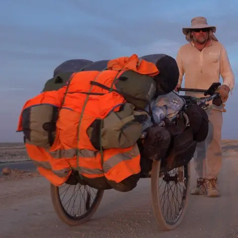Karl Bushby Karl Bushby wearing a dusty white top, pants, black sunglasses and a wide brimmed beige hat. He is pushing a cart, which contains his belongings covered in an orange hi-vis fabric, along a dusty road.