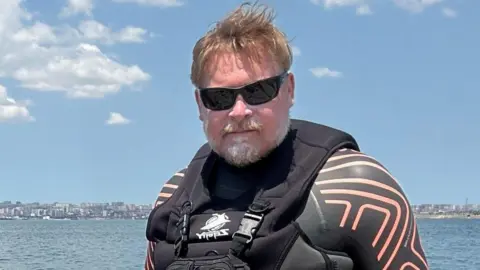 Karl Bushby Karl Bushby has short blonde hair and a grey beard. He is wearing black sunglasses and a black wetsuit with geometric orange-coloured lines on the shoulder and sleeves. The Caspian Sea is in the background and looks calm and bright on a sunny day. The sky is blue with scattered white fluffy clouds in the top left corner.