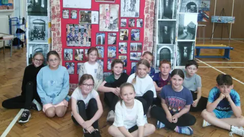 Quay Academy Twelve primary school children sitting on a wooden herringbone floor in front of large display of artwork.
