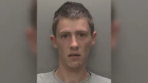 Humberside Police A police headshot of a man wearing a light grey t-shirt with short brown hair looking at the camera. He is stood in front of a grey wall.