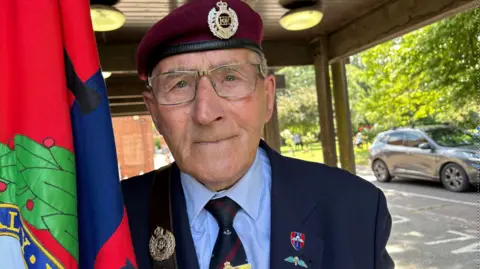BBC News A man wearing a burgundy beret with glasses in a navy suit, looking into the camera. He is holding a blue, red and green regimental flag.