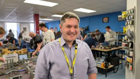 BBC News / Josh Gorroño Chapman Adam Stephenson looks into the lens smiling. He is wearing a checked lilac shirt with a yellow volunteer lanyard around his neck.