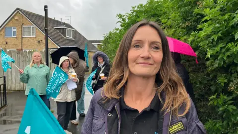 Amanda White/BBC A woman with long brown hair smiling at the camera. She is wearing a dark Barbour jacket. Behind her are several people are holding teal-coloured flags with white writing, wearing raincoats and using umbrellas. There are residential houses behind a wooden fence in the background.