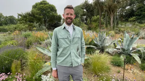 BBC/Jo Makel A man wearing a white shirt, mint green jacket and striped trousers stands amongst a garden featuring spiked plants and colourful flowers. He has short cropped brown hair, a beard and is wearing glasses.