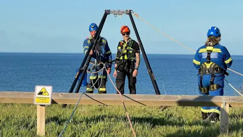 Bridlington Coastguard Rescue Team Two men wearing helmets and ropes standing at the edge of a cliff. One man is dressed in a blue HM Coastguard uniform and the other is in black police uniform. The sea is behind them and a sign says "Dangerous cliffs".