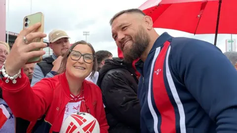 BBC/Matt Dean A crowd of rugby fans with player Jared Waerea-Hargreaves. A woman at the front of the crowd is holding a mobile phone up in the air on the left and smiling. She has oversized black-framed spectacles on her face and is wearing a red bomber jacket over a red and white t-shirt. Her brown hair is tied up in a ponytail. She is holding a red and white rugby ball with the right hand and is standing next to a rugby player, who is smiling at towards the mobile phone. He is wearing a blue long-sleeved top with a red vertical stripe along the right of his chest.