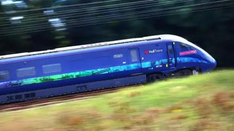 Hull Trains A Hull Trains train in dark blue named Land of Green Ginger with images of the city on a strip down the side speeding along a railway line with the grass in front of it and the overhead wires blurred