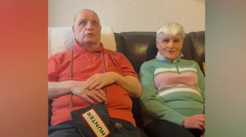 Lynne and Trevor Man with red polo shirt and dark trousers has a bag hanging from his neck and placed on his lap with Hunter written on it. He is sat on a brown sofa with a woman with a patterned green top and dark trousers. There is a beige wall. 