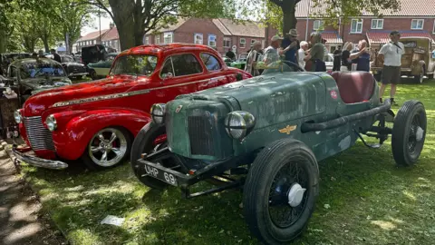 A red vintage car is parked alongside a green vehicle on a patch of grass in the shade of a tree.