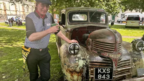 A man wearing a flat cap, polo shirt and dark jeans stands next to a rusty looking pickup truck