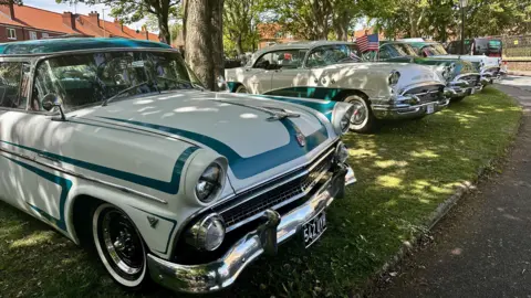 Four vintage cars, in cream and green colours, line a path surrounded by trees