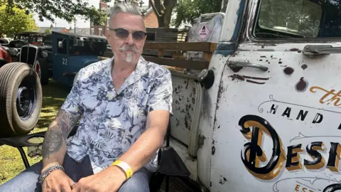 A man with grey hair, sunglasses and a beard sits next to a white truck. He is wearing dark jeans and a white Hawaiian style shirt with flowers on it.