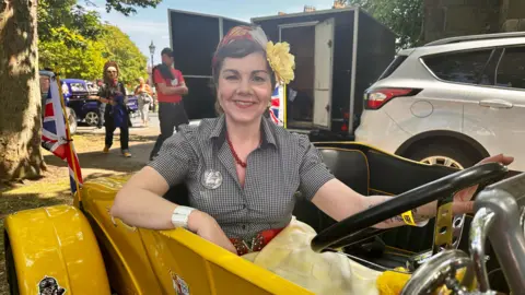 BBC A woman with dark hair, a headscarf and large yellow flower smiles as she sits in black and yellow car. She is wearing a black check dress with a red necklace and red belt.
