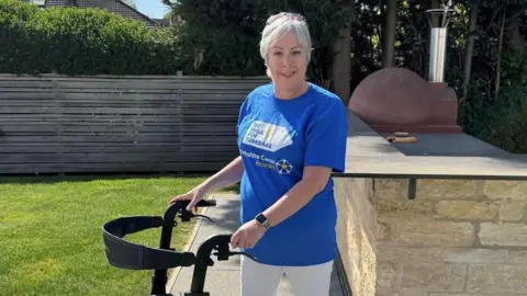 Yorkshire Cancer Research Louise Hatfield wearing a blue Yorkshire Cancer Care t-shirt, white trousers and short white hair. She is standing with a walking frame in a garden with grass, paving, a fence, trees and a brick bar area. She is wearing a watch. She is smiling.
