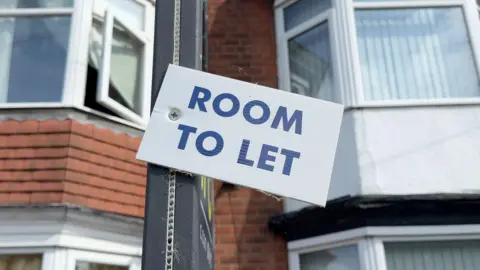 BBC Room To Let sign in blue capital letters on a white background in front of terraced housing in Hull
