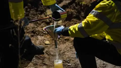 Getty Images Two workmen in hi-vis jackets sift water into a bottle through a funnel. They appear to be working on a sandy surface.