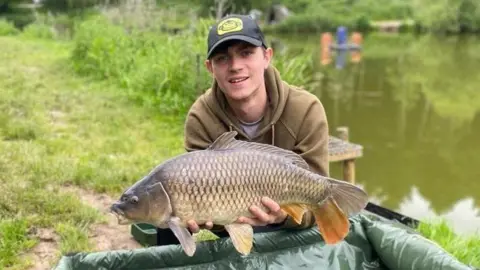 Family Handout A man wearing a black baseball cap and brown hoodie is crouched on a patch of grass close to a large area of water. He is holding a large fish.