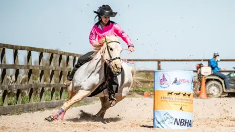 Supplied A girl wearing a cowboy hat, pink shirt and black trousers rides a grey horse at speed around and blue and yellow barrel, in a sandy arena surrounded by a fence