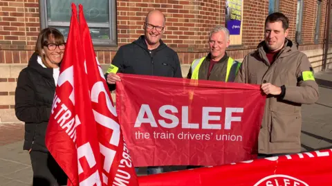 Four people standing on a street corner holding red banners and flags bearing the words "Aslef, the train drivers' union". The three men and a woman are smiling and standing in front of a large red brick building.