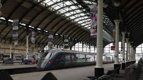 BBC A blue train at a large, covered railway platform with high arched ceilings made of glass and metal beams. White pillars support the structure, and banners hang from the ceiling. The platform includes benches for passengers.