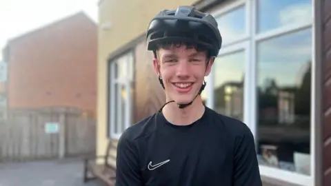 Sarah Sanderson/BBC A landscape head and shoulders shot of a teenager in a black top with a white tick on it wearing a black helmet. He is smiling into the camera in front of a house.