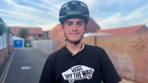 Sarah Sanderson/BBC A teenage boy is wearing a black top with a white skateboard logo which reads: 'VANS OFF THE WALL'. He is wearing a dark green helmet and smiling into the camera. You can see a path behind him leading to what looks like a garage and on the right is a red brick wall.