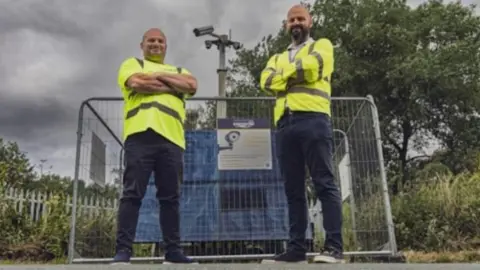 East Riding of Yorkshire Council Two men stand posing for the camera with their arms folded, wearing hi-vis jackets, jeans and trainers. They are stood in front of the cameras, which have metal fences around them.