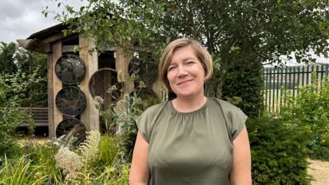 BBC Sarah Weichardt smiles directly at the camera. She has mousey blonde hair that is just past her ears. She wears a green blouse and is pictured in front of a wooden shed with circular metal holes patterned with bird designs on. There is lush greenery behind her, and a variety of plants are growing wildly throughout. You can see a glimpse of a green metal fence to her right, with houses and a field behind this.
