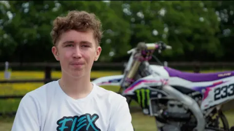 Yorkshire Air Ambulance Boy with short brown hair and white t-shirt. There is a white and purple motorbike behind him and trees and grass.