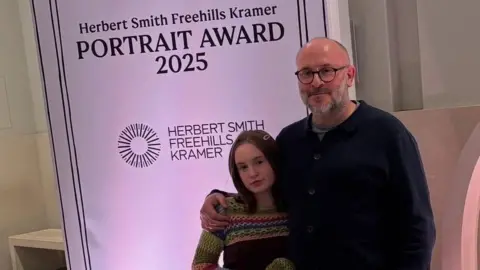 Submitted Girl with colourful dress and long brown hair stands in front of a banner that reads "Herbert Smith Freehills Kramer Portrait Award 2025". She is stood with a man with glasses and a beard with a dark jumper.