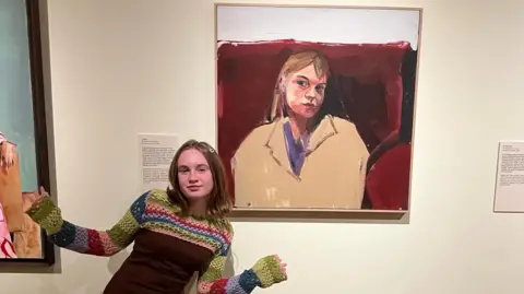 Submitted Girl with colourful dress stands in front of a portrait of her on a wall. She has long brown hair.
