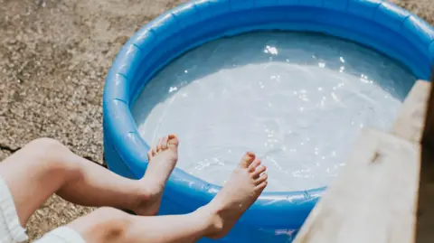 Getty Images A pair of feet rest against a full paddling pool during hot sunshine.