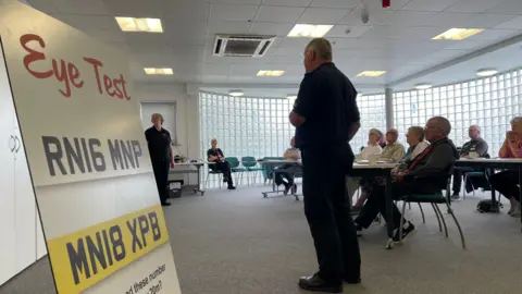 A group of people are sitting behind desks looking at a presentation screen. In the foreground is a board saying "eye test". It also shows two number plates and instructions to try to read the plates from 20m away.