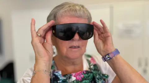 A woman with grey hair is trying on a pair of large black glasses. The lenses have black speckles on them.