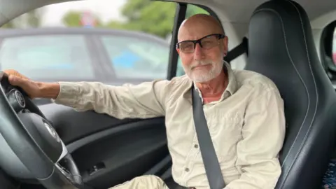 BBC A man sits in the driving seat of a car, with his right hand on the steering wheel and his seat belt on. He has a bald head and a white beard. He is wearing glasses and smiling at the camera. He is wearing a cream shirt.