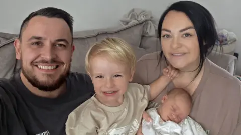 Manchester University NHS Foundation Trust Man with short dark hair and beard with a dark top with a child with blonde hair and a beige top, a woman with brown hair and a brown top and a baby wrapped in white. They are sitting on a sofa.