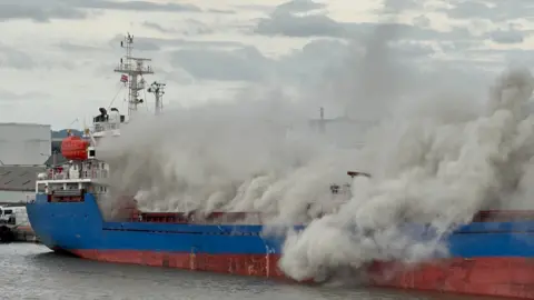 BBC A blue and red cargo ship has thick smoke billowing out of it