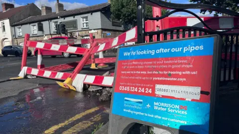 BBC A water leak on a busy main road which has been cordoned off with a Yorkshire Water sign stating We're looking after our pipes
