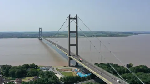 PA Media An aerial shot of a large bridge spanning a wide brown river, with tall towers and cables holding it up. Cars are travelling across the bridge. There are houses, trees and a small construction site near the bridge's base in the foreground of the photo. 
