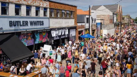 A large crowd in the sunshine in Humber Street, Hull. A series of converted warehouses, now home to restaurants and shops, stand on the far side of the street. To the left is a restaurant with a blue sign reading "Butler Whites" and shutters painted with colourful graffiti.