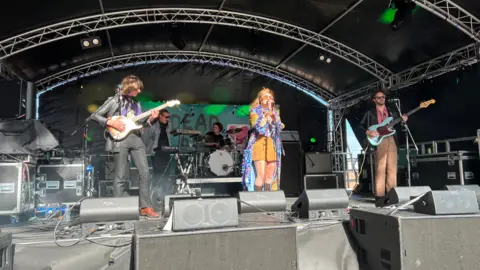 BBC A female signer wearing a flowing, purple, blue and yellow dress leads a four-piece band, including two guitarists and a drummer, on a stage with black tarpaulin-style walls and roof at Humber Street Sesh in Hull