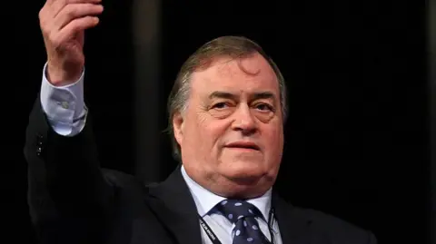 Getty Images John Prescott pictured wearing a black suit jacket, a light blue shirt and a dark blue tie with light blue spots. He has short hair and is holding up his right arm, pointing it to the sky.